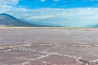 some mountains and a pink lake surrounded by rocks and dirt in front of a blue sky with clouds