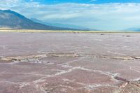 some mountains and a pink lake surrounded by rocks and dirt in front of a blue sky with clouds