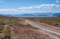 a dirt road passing through the desert under a partly cloudy sky with mountains behind it