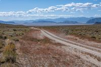 a dirt road passing through the desert under a partly cloudy sky with mountains behind it