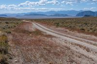 a dirt road passing through the desert under a partly cloudy sky with mountains behind it