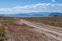 a dirt road passing through the desert under a partly cloudy sky with mountains behind it