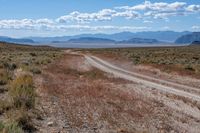 a dirt road passing through the desert under a partly cloudy sky with mountains behind it