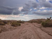 a dirt road in the middle of an arid area under clouds and sun set over mountains