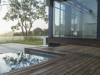 a pool with a wooden deck, a patio and a large glass window with trees in the background