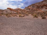 a couple of red rocks in the desert and some bushes and dirt and a cloudy sky