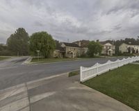 the view down a street that includes homes and lawn areas with white picketers, trees, and clouds