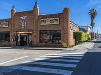 a brick building sits on the corner of an empty street with crosswalks and palm trees