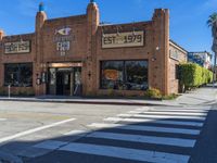 a brick building sits on the corner of an empty street with crosswalks and palm trees