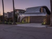 a modern home with a curved roof and exterior in the evening sun light, sitting in a driveway between palm trees