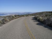 the empty road is surrounded by brush and sand and mountains in the distance in the desert