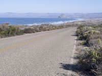 an empty road with two lanes stretching along a shoreline in front of the ocean and mountains