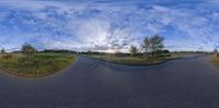 a road near some grass and bushes with the sun in the background and clouds overhead