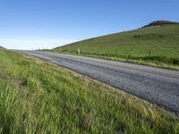 an empty road with a single lane through tall grass and a hill on the other side