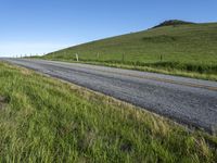 an empty road with a single lane through tall grass and a hill on the other side