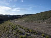 a dirt road winding into a small hill side village surrounded by grass, shrubs and blue sky