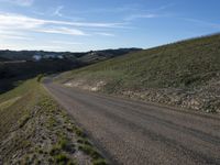 a dirt road winding into a small hill side village surrounded by grass, shrubs and blue sky