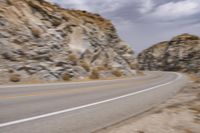 a motorcycle traveling down a street next to a mountain side of a cliff and an empty road in the middle
