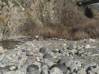 a river flowing under a bridge next to a rocky hillside side covered in vegetation and small boulders