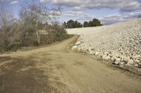 a graveled dirt road on a sunny day near a field of rocks and trees