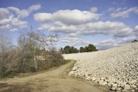 a graveled dirt road on a sunny day near a field of rocks and trees