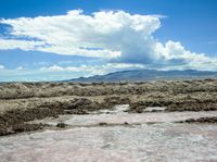 a man that is standing in some dirt by some water, clouds and mountains behind him