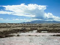 a man that is standing in some dirt by some water, clouds and mountains behind him