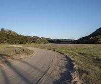 a dirt road in front of a lush green hillside side forest with a clear blue sky