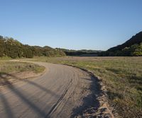 a dirt road in front of a lush green hillside side forest with a clear blue sky