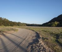 a dirt road in front of a lush green hillside side forest with a clear blue sky