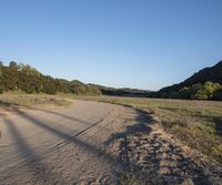 a dirt road in front of a lush green hillside side forest with a clear blue sky