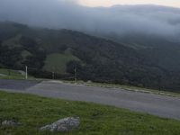 a road that goes up a hill and has green mountains behind it with clouds overhead