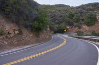 a curve road going through a rocky area with trees on both sides and a yellow painted sign that reads,