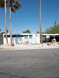 a white house sits on the corner of the street with two palm trees and some rocks