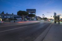 the lights of vehicles and some buildings are on a city street at twilight with a neon sign and street signs