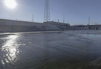 a body of water with sun reflecting in the water and power towers behind it with electricity wires