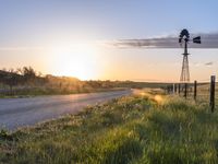 a rural countryside view with windmill and sun setting in the horizon with green grass, fence, and a dirt road