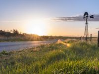 a rural countryside view with windmill and sun setting in the horizon with green grass, fence, and a dirt road