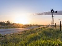 a rural countryside view with windmill and sun setting in the horizon with green grass, fence, and a dirt road