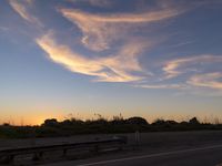 a long bench is beside an empty field with a sky view at sunset behind it