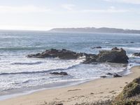 the surfer is riding on the sand with his surfboard near some rocks and the ocean