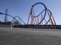 a roller coaster sits in front of a building and is blue with orange accents and has two red steel bars on the sides of it