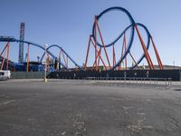 a roller coaster sits in front of a building and is blue with orange accents and has two red steel bars on the sides of it