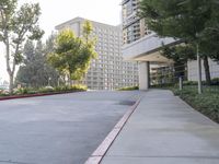 a paved sidewalk and side walk leading to buildings and trees next to the sidewalk, with parking spaces and parked cars