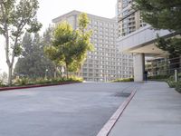 a paved sidewalk and side walk leading to buildings and trees next to the sidewalk, with parking spaces and parked cars