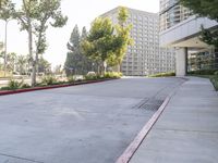 a paved sidewalk and side walk leading to buildings and trees next to the sidewalk, with parking spaces and parked cars