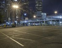 an empty lot next to a lot with parking spaces and tall buildings at night time