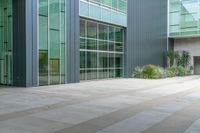 the concreteed walkway is lined with glass windows and a bench outside a building that is surrounded by greenery