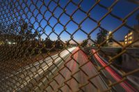 blurred street lights are seen in the distance through a chain link fence overpassed with a highway