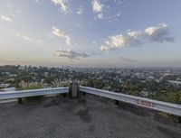 a parking lot with a concrete bench and metal railings over looking the city below a big blue cloudy sky